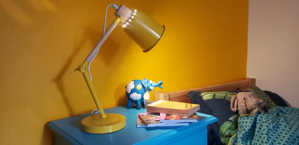 tirelire éléphant en céramique bleue posée sur la table de nuit d'un enfant, avec une lampe jaune et le mur derrière jaune.